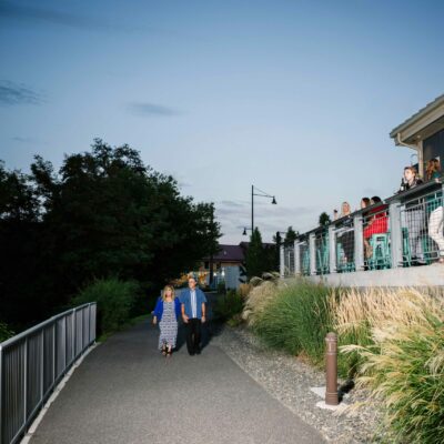 A couple strolls along the waterfront path at Columbia Gardens past groups visiting and sipping wine at Muret-Gaston Winery's tasting room patio.